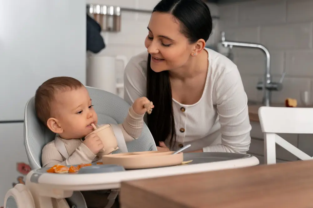 Mãe sorrindo para o bebê, que bebe de um copinho no cadeirão, com pratinho de silicone na bandeja