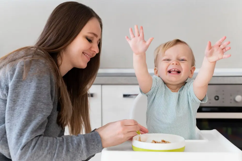 Mãe sorrindo enquanto o bebê, no cadeirão, levanta os bracinhos e ri